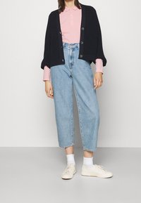 Mörkstickad cardigan med knapprader, rosa skjorta med krage, ljusblå högmidjade jeans och beige sneakers med vita strumpor.