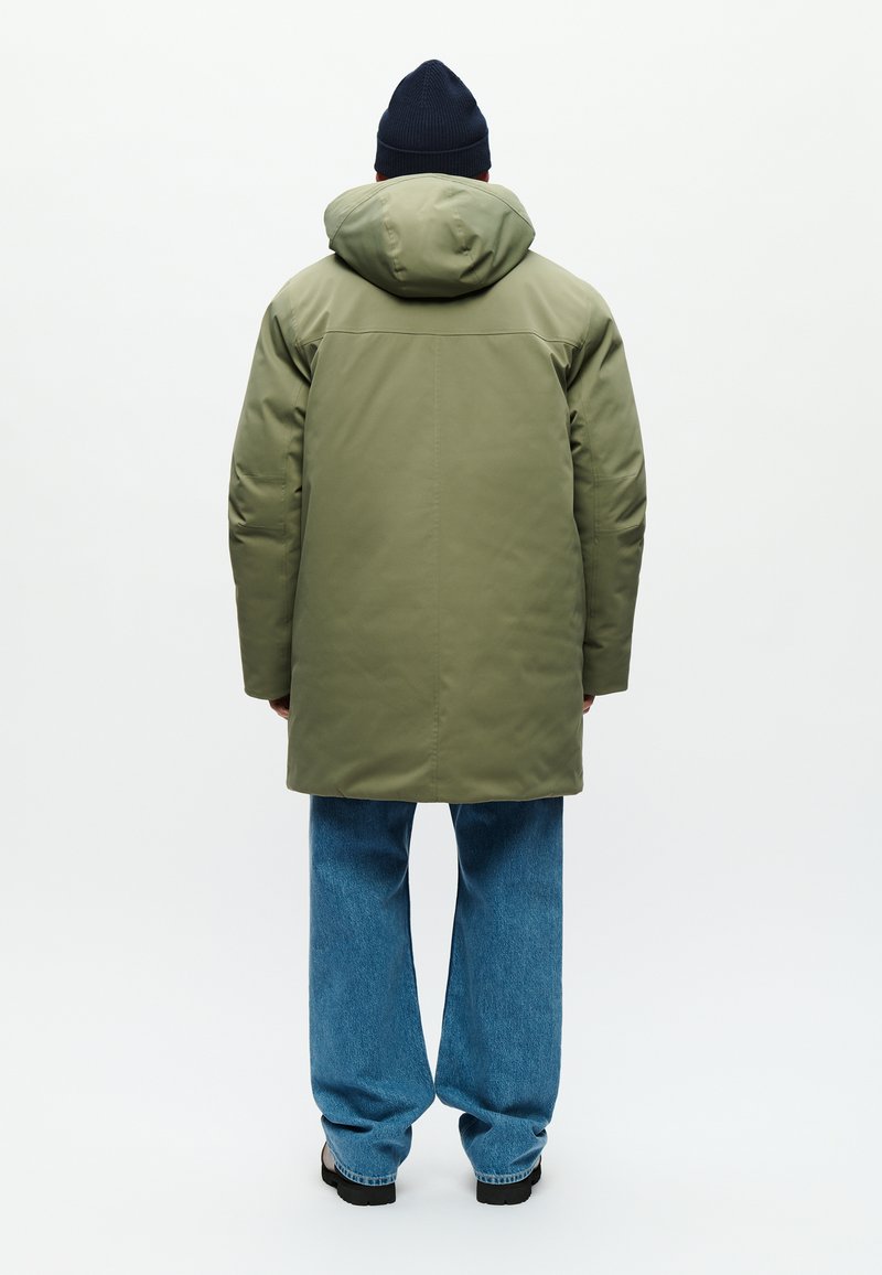 Parka aislante de color verde oliva con capucha, que presenta un corte recto y detalle en la costura trasera, combinada con unos vaqueros de pierna ancha de color azul claro.