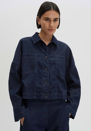 Person mit kurzen dunklen Haaren, die eine dunkelblaue Jeansjacke mit Knöpfen und passende Hose vor einem schlichten, hellen Hintergrund trägt.