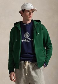 Giovane uomo con cappellino bianco, felpa verde con zip, felpa blu navy con logo e pantaloni della tuta grigi, che guarda di lato su sfondo neutro.