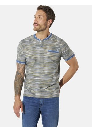 Mann steht, blickt nach links, trägt ein kurzärmeliges, gestreiftes Hemd mit blauen Abschlüssen und Tasche, blaue Jeans und hat Tätowierungen am linken Arm.