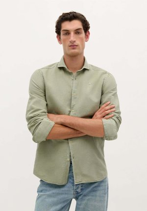 Light green long-sleeve shirt with a button-down collar, featuring a subtle texture and a small logo on the chest. Paired with blue jeans.
