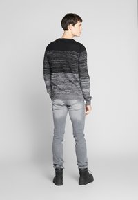 Čierny a šedý pruhovaný pletený sveter s rebrovaným výstrihom, v kombinácii so šedými úzkymi džínmi a čiernymi členkovými topánkami. Model otočený chrbtom, neutrálne pozadie.