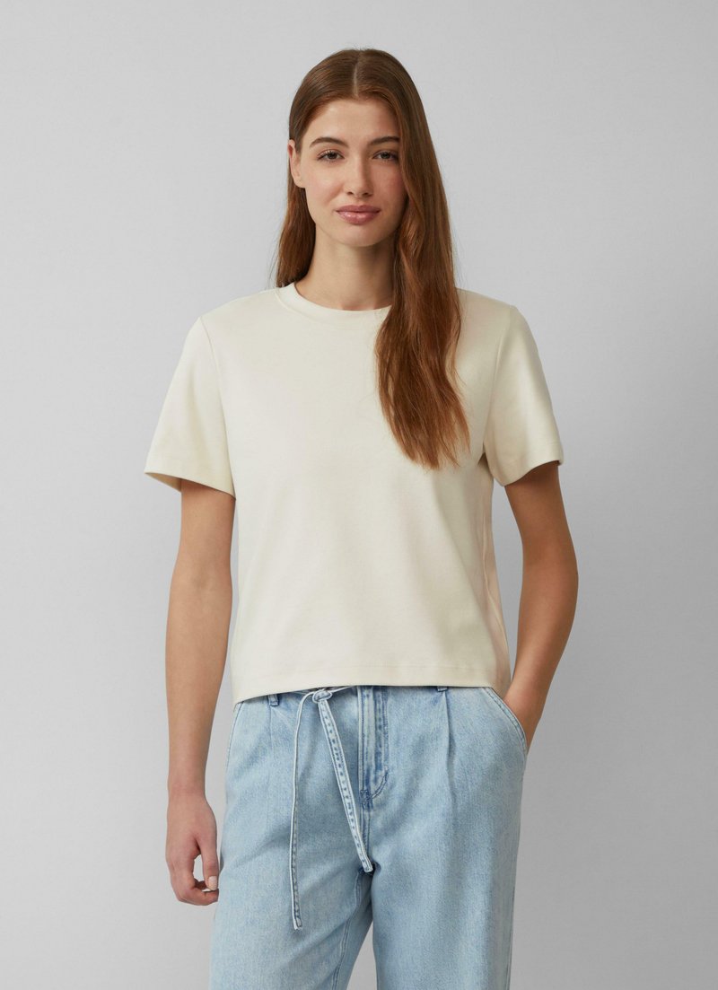 Kremowy t-shirt z krótkim rękawem i okrągłym dekoltem, wykonany z gładkiego materiału, w połączeniu z jasnoniebieskimi spodniami dżinsowymi na sznurki, o luźnym kroju.