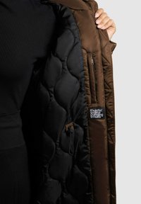 Interior de chaqueta tipo puffer marrón con forro acolchado negro, con un bolsillo con cremallera y un parche con el logotipo. Textura de tela suave e insulated.