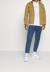 Sandfärgad manchesterskjorta med två bröstfickor över en vit t-shirt med tryck, kombinerad med korta blå jeans och vita sneakers.