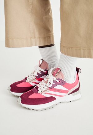 Osoba w beżowych spodniach, białych prążkowanych skarpetach i różowo-fioletowo-białych sportowych butach na sznurowadła stojąca na białej powierzchni.