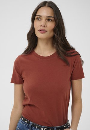 Giovane donna con capelli lunghi e scuri che indossa una maglietta a maniche corte color ruggine e jeans blu con una cintura nera borchiata.