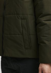 Zielona oliwkowa kurtka ocieplana z zamkiem błyskawicznym i pionową kieszenią boczną. Materiał ma teksturowane wykończenie z widocznymi szwami.