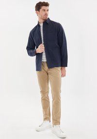 Camicia blu navy con taschino, camicia interna a righe grigie e bianche, pantaloni beige e sneakers bianche. Tessuto morbido, vestibilità sartoriale.