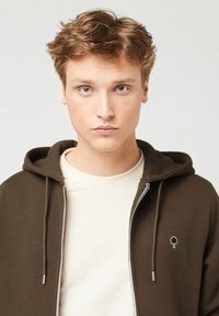Felpa marrone con zip, foderata in grigio e con coulisse, dotata di un piccolo logo ad albero. Indossata sopra una maglietta chiara.