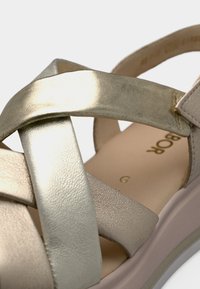 Χρυσό μεταλλικό σανδάλι με λουράκια και υφή, με ανυψωμένη, απαλό ροζ σόλα και ραμμένα διακοσμητικά στοιχεία για επιπλέον λεπτομέρεια.