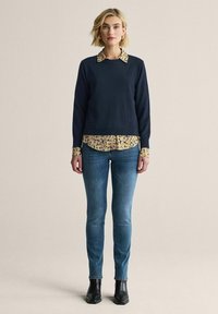 Marinblå sweatshirt med rundad halsringning, lager-på-lager över en blommönstrad ljusgul skjorta. Ett par blå tajta jeans och svarta ankelboots.