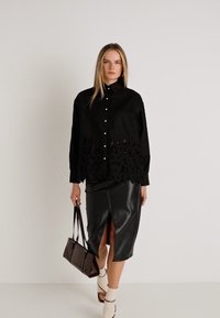 Camisa preta de botão com detalhes em renda na barra, combinada com uma saia de couro preta com uma abertura lateral. Mules brancas de salto e uma bolsa castanha.