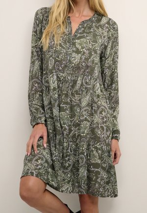 Frau trägt knielanges, langärmliges olivgrünes Kleid mit weißem und schwarzem Paisley-Muster, steht vor einfarbigem Hintergrund.