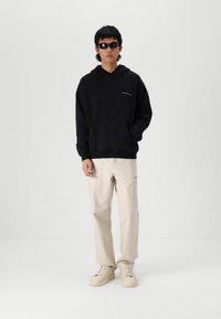 Hanorac negru cu glugă, cu un buzunar frontal, asociat cu pantaloni cargo bej deschis și adidași crem. Accesoriile includ ochelari de soare închiși la culoare.