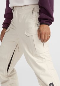 Beige cargo pants featuring multiple pockets, zipper details, and a tapered leg design. Smooth texture with subtle sheen.