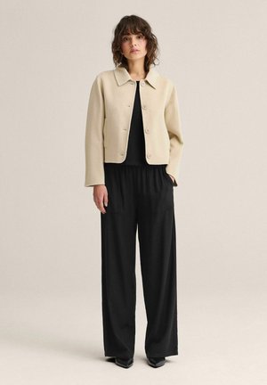 Kvinde stående iført beige cropped jakke med knapper, sort top, sorte bukser med vide ben og sorte spidse sko mod en ensfarvet baggrund.