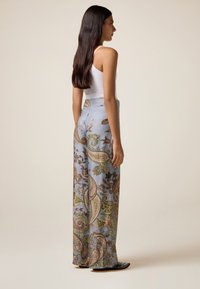 Donna in profilo che indossa una canotta bianca senza maniche, pantaloni a vita alta e larghi con stampa paisley, e scarpe nere con borchie.