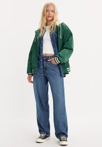 Zielona wodoodporna kurtka z białymi akcentami, nałożona na dżinsową koszulę i niebieskie szerokie spodnie. Czarne trampki dopełniają stylizację.