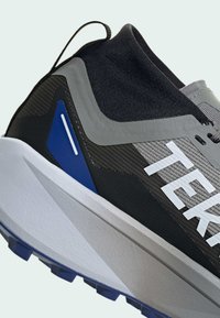 Scarpa sportiva grigia e nera con tomaia in rete, logo bianco, dettagli testurizzati e una spessa suola bianca con battistrada blu e nero.