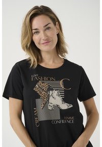 T-shirt noir avec un design graphique comprenant des bottes en imprimé beige et léopard, des accents de texte en doré, un col rond et des manches courtes.