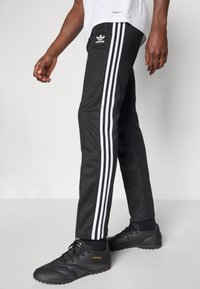 Černé sportovní kalhoty se třemi bílými pruhy po stranách, na levém stehně s logem Adidas, doplněné o černé boty.
