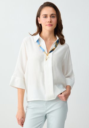 Femme aux cheveux bruns mi-longs portant une chemise blanche ample et un pantalon clair, posant avec la main dans la poche devant un fond uni.