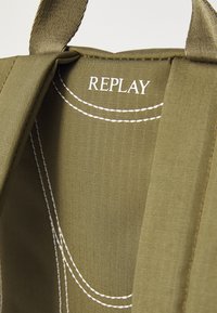 Grønt stoff med en strukturert overflate. Har hvite sømmer og ordet "REPLAY" trykt i hvitt på innsiden.