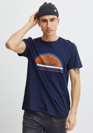 Férfi sötétkék "Blend Denim Original Built in Blue" feliratú pólóban és sötét kötött sapkában, sima világos háttér előtt pózol.