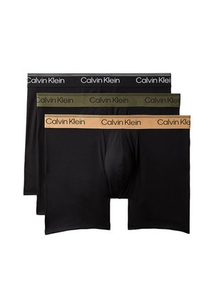 Skjorteløs mand justerer sorte Calvin Klein boxer shorts, stående barfodet mod en ensfarvet grå baggrund.