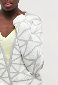 White cardigan with gray geometric patterns, knitted texture, and open front design. Worn over a light yellow v-neck top.