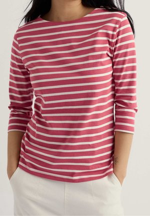 Femme portant un haut à manches longues rayé rose et blanc avec des manches trois-quarts et un pantalon blanc, les mains dans les poches.