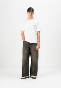 Mladenič stoji oblečen v črno kapo, belo grafično majico, široke črne jeans hlače in bele športne copate proti enotni beli podlagi.
