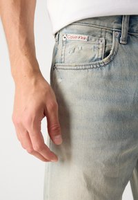 Vaqueros de mezclilla de color azul claro con detalles desgastados, que presentan una etiqueta de Calvin Klein, un botón plateado y acentos de costura.