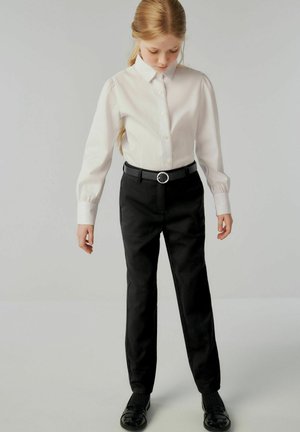 Witte langemouwen shirt met knoopsluiting, zwarte riem met een cirkelvormige gesp en zwarte, stijlvolle broek. Zwarte schoenen met textuurdetail.