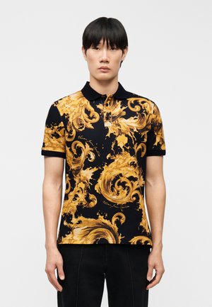 Noor mees, kes kannab musta polo-särki keerulise kuldse barokkstiilis mustriga, seisab käed lõdvestunud asendis täiesti ühevärvilise tausta ees.