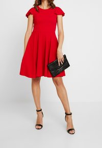 Czerwona sukienka z krótkimi falbanowymi rękawami, dopasowanym gorsetem i rozkloszowaną spódnicą. Do kompletu czarna, teksturowana kopertówka i czarne sandały na paski.