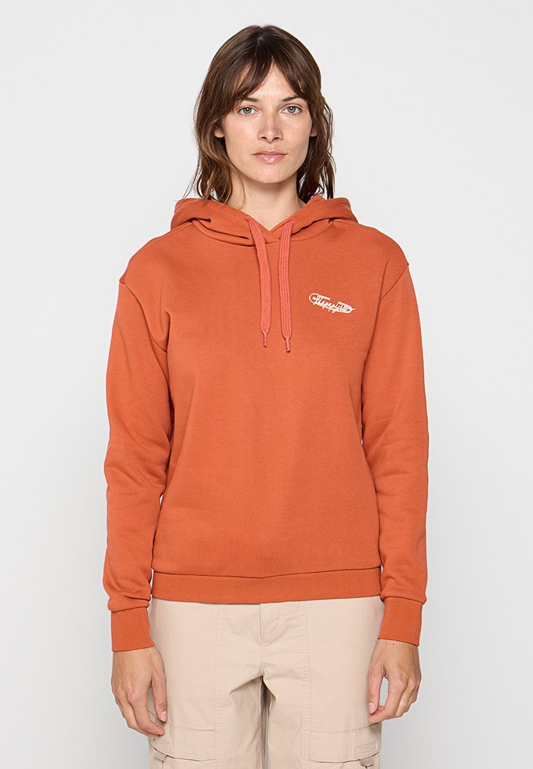 Kobieta ubrana w rudobrązową bluzę z kapturem z małym białym logo na lewej piersi oraz beżowe spodnie cargo, stojąca na tle jasnym.