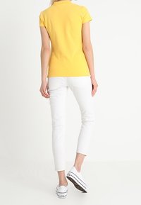 Personne portant un polo jaune à manches courtes, un pantalon blanc ajusté et des baskets blanches, se tenant contre un fond blanc uni.