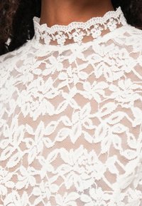 Fehér csipkeblúz bonyolult virágmintákkal, magas csipke gallérral és félig átlátszó, textúrált felülettel.