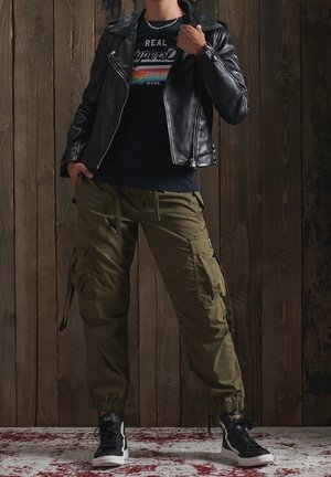 Osoba ubrana w czarną skórzaną kurtkę, czarną grafikową koszulkę, oliwkowe spodnie cargo i czarne wysokie trampki, stoi w pomieszczeniu.