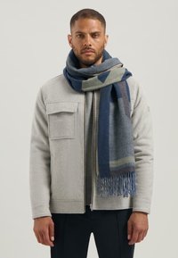 Grijs fleece jacket met een rits, voorzien van twee vo pockets. Een blauw geruite sjaal met franjes hangt om de nek.