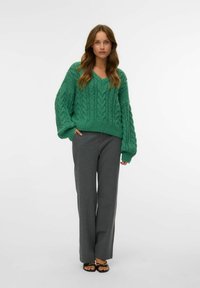 Pull à mailles en vert avec un col en V, associé à un pantalon large à rayures grises. Le pull présente des manches surdimensionnées et texturées.