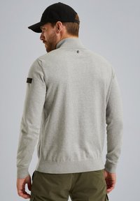 Jasnoszary sweter typu pulower w teksturowanym splocie, z podniesionym kołnierzem i naszywką z logo po boku. Rękawy i dół są wykończone ściągaczem.