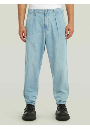 Jean bleu clair plissé et coupe ample, porté avec des chaussures noires à lacets et un haut blanc, présenté de la taille aux pieds sur un fond neutre.
