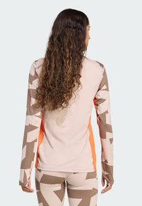 Langärmlig sportshirt in lichtroze met bruine geometrische patronen en oranje accenten. Heeft een nauwsluitende pasvorm en verlengde manchetten.