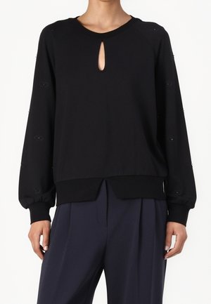 Mujer que lleva una blusa negra de manga larga con escote en forma de ojo de cerradura y detalles de botones en las mangas, combinada con pantalones oscuros plisados.