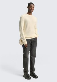 Crème geribbelde trui, donkere jeans en zwarte enkellaarsjes. Het model houdt een kleine crèmekleurige tas vast. Eenvoudig ontwerp met textuurstof en neutrale kleuren.