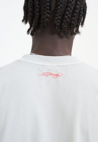 Světle šedé tričko s kulatým výstřihem, měkkou texturou a vyšívaným červeným logem "Ed Hardy" na zadní straně blízko límce.